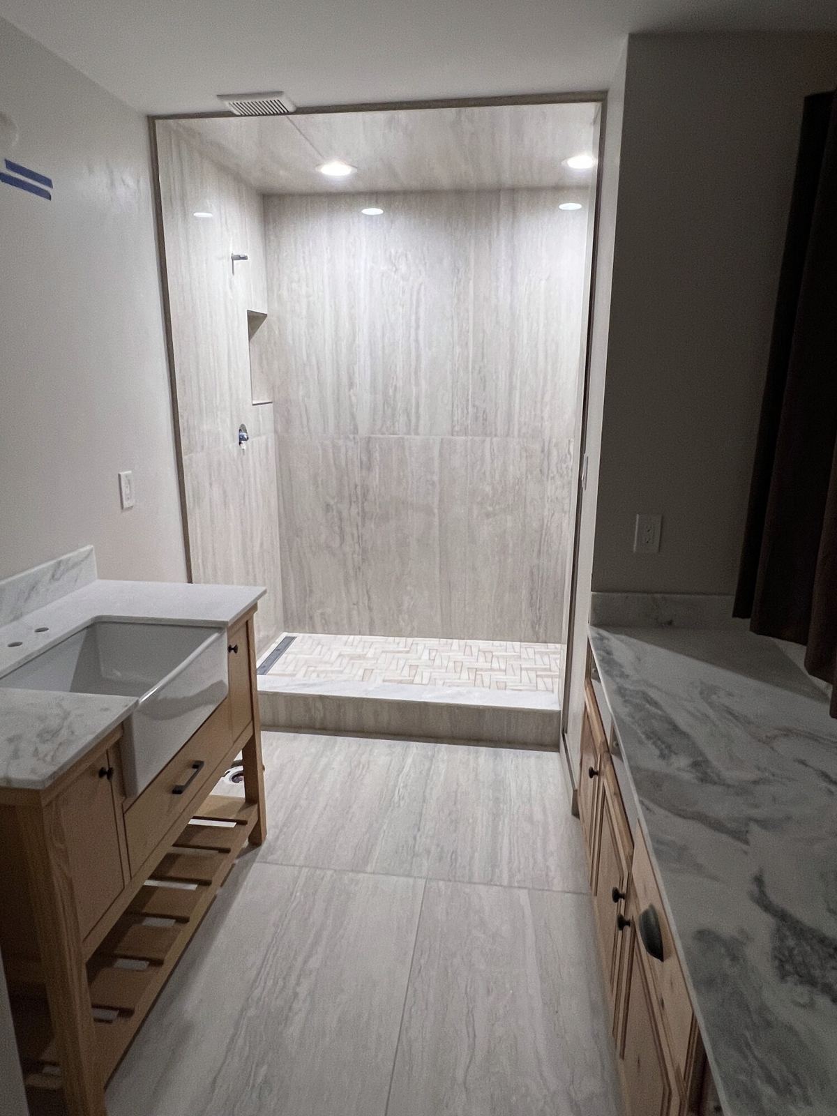 Bathroom with a walk-in shower at the end, wood-look tile floor and stone vanity tops on both sides